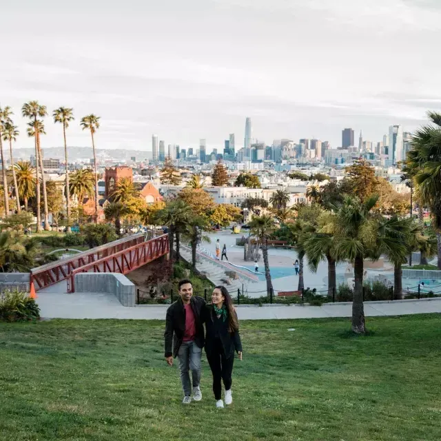 Una coppia cammina verso la telecamera con Dolores Park e lo skyline di 贝博体彩app dietro di loro.